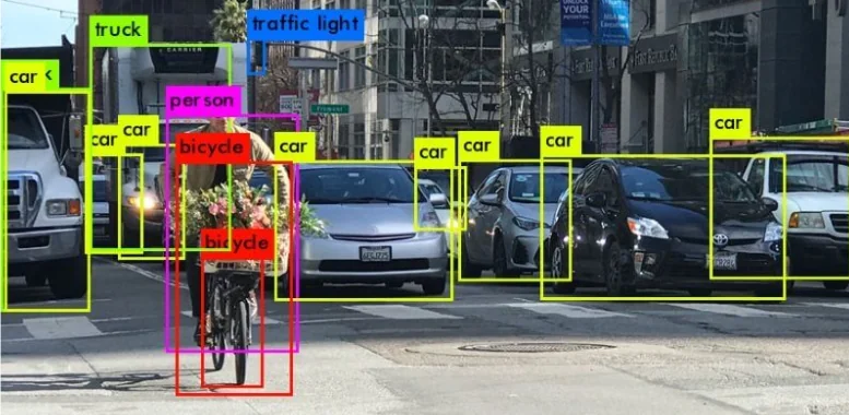 Object-detection demo photo of a city street: a person cycling with flowers on the bike surrounded by traffic, with coloured bounding boxes labelled person, bicycle, car, truck and traffic light